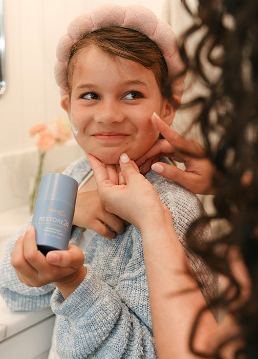 Child holding a RESTORE24 bottle while an adult woman applies skin care to the child's face.