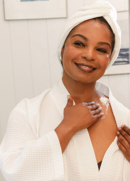 Woman applying RESTORE24 cream to her shoulder with a towel on her head in a bathroom setting